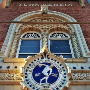 May include: A vintage brick building with a white archway and a large, ornate crest. The crest features a blue and white design with the words "American Turnerbund" and "Sound Mind Sound Body" in a circle around a figure throwing a discus. The year 1854 is inscribed below the figure. The building has the words "Turn Verein" above the archway.
