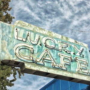 May include: A faded blue and white sign with the words "Lucky Cafe" in neon letters. The sign is hanging from a building and has a weathered, vintage look.