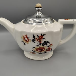 Könnte beinhalten: Weiße Keramik-Teekanne mit silbernem Deckel und floralem Design in Rot, Gelb und Schwarz. Die Teekanne hat einen gebogenen Ausguss und einen Griff mit einem kleinen floralen Akzent. Ein dekoratives Stück zum Servieren von Tee.