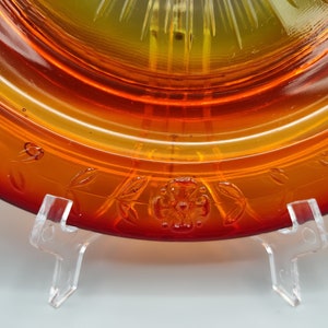 May include: Close-up of an amber-colored glass plate with a floral and leaf design. The plate has a gradient of colors, transitioning from a deep red to a yellow center. The plate is displayed on a clear stand.
