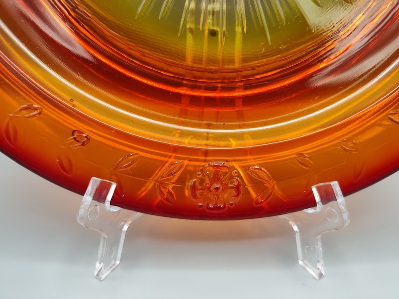 May include: Close-up of an amber-colored glass plate with a floral and leaf design. The plate has a gradient of colors, transitioning from a deep red to a yellow center. The plate is displayed on a clear stand.