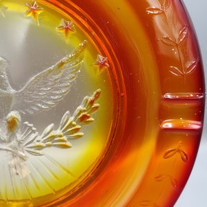 May include: An iridescent orange and yellow glass ashtray with a detailed embossed design. The ashtray features a central image of a bird with outstretched wings, surrounded by stars and floral accents. The rim has a leaf pattern.