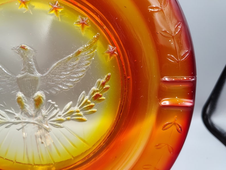 May include: An iridescent orange and yellow glass ashtray with a detailed embossed design. The ashtray features a central image of a bird with outstretched wings, surrounded by stars and floral accents. The rim has a leaf pattern.