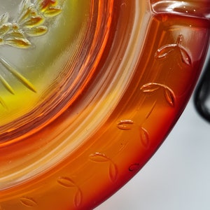 May include: Close-up of a decorative glass dish with a vibrant orange and yellow color gradient. The rim features embossed leaf designs. The center has a detailed embossed design of a wreath and sunburst.