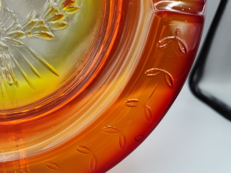 May include: Close-up of a decorative glass dish with a vibrant orange and yellow color gradient. The rim features embossed leaf designs. The center has a detailed embossed design of a wreath and sunburst.