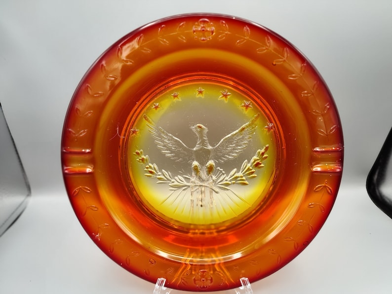 May include: An orange glass ashtray with a central design featuring a detailed eagle with outstretched wings, surrounded by stars and olive branches. The rim has leaf and flower patterns, and it has three cigarette rests.