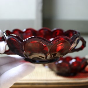 Könnte beinhalten: Eine rubinrote Glasschale mit gewelltem Rand und zwei kleinen Griffen. Die Schale hat eine glänzende Oberfläche und wird in einer behandschuhten Hand gehalten. Wahrscheinlich Vintage oder Antik.