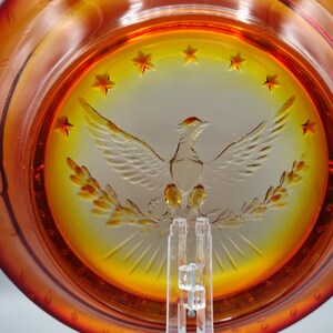 May include: Amber-colored glass plate with a central design of a spread-winged bird, surrounded by stars. The plate has a decorative rim and is displayed on a clear stand. The design is etched into the glass.