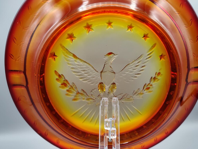 May include: Amber-colored glass plate with a central design of a spread-winged bird, surrounded by stars. The plate has a decorative rim and is displayed on a clear stand. The design is etched into the glass.