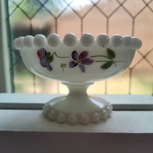 May include: White milk glass pedestal bowl with a beaded rim and base. The bowl features hand-painted purple and white violets with green leaves. The bowl is sitting on a white windowsill.