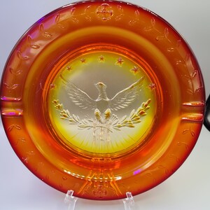 May include: An orange and yellow glass ashtray with a central embossed design featuring a dove, olive branches, and stars. The rim has a decorative leaf pattern. The ashtray is displayed on a clear stand.