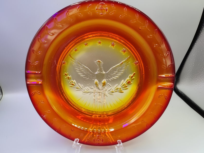May include: An orange and yellow glass ashtray with a central embossed design featuring a dove, olive branches, and stars. The rim has a decorative leaf pattern. The ashtray is displayed on a clear stand.