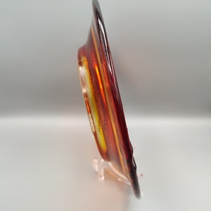 May include: A decorative glass plate with a vibrant color scheme of red, orange, and yellow. The plate is displayed on a clear stand, showcasing its unique design and artistic flair. The plate's colors create a warm and inviting aesthetic.