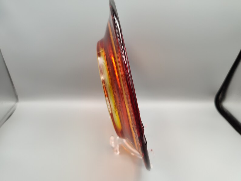 May include: A decorative glass plate with a vibrant color scheme of red, orange, and yellow. The plate is displayed on a clear stand, showcasing its unique design and artistic flair. The plate's colors create a warm and inviting aesthetic.