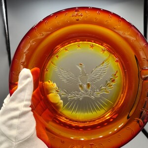 May include: Amber-colored glass ashtray with a central etched design of an eagle with outstretched wings, surrounded by stars and laurel branches. The rim features a decorative leaf pattern. A classic piece of home decor.
