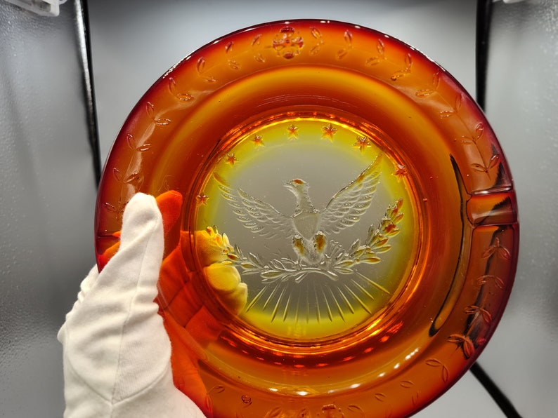 May include: Amber-colored glass ashtray with a central etched design of an eagle with outstretched wings, surrounded by stars and laurel branches. The rim features a decorative leaf pattern. A classic piece of home decor.