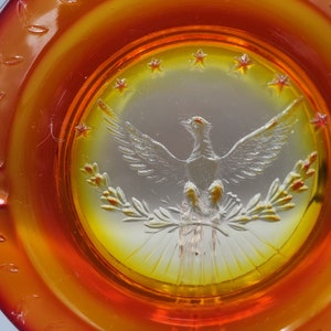 May include: An amber-colored glass ashtray with a central design featuring a detailed eagle with outstretched wings, surrounded by stars and laurel branches. The ashtray has a circular shape with a decorative leaf pattern around the rim.