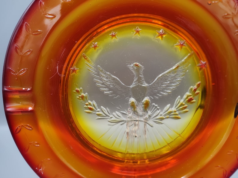 May include: An amber-colored glass ashtray with a central design featuring a detailed eagle with outstretched wings, surrounded by stars and laurel branches. The ashtray has a circular shape with a decorative leaf pattern around the rim.
