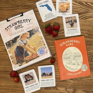 May include: An educational literature unit titled "Strawberry Girl" with a book and flashcards about Florida. The book is orange with the title and author's name. Flashcards show Florida's state fruit, hogs, and a horse. Fresh strawberries are scattered around.
