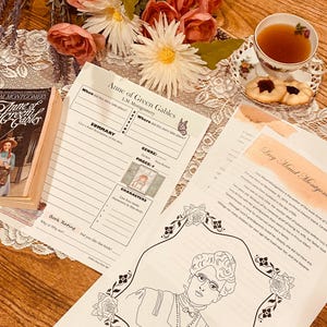 Può includere: Una copia aperta di "Anne of Green Gables" di L.M. Montgomery è accanto a fogli di lavoro e una tazza da tè con biscotti. La copertina del libro mostra una donna in abito e cappello. Fiori e pizzo completano la scena.
