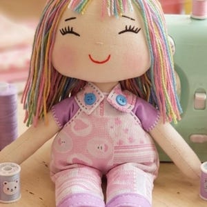 Puede incluir: Una muñeca de tela con cabello de hilo multicolor, ojos cerrados bordados y una sonrisa cosida en rojo. La muñeca viste una camisa morada clara bajo un overol rosa con patrones de corazones y círculos, acentuado con dos pequeños botones azules en el cuello.