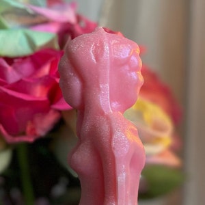 Könnte beinhalten: Eine rosa, weibliche Kerze mit strukturierter Oberfläche steht auf einer rosa Stoffoberfläche mit zwei rosa Rosen. Im Hintergrund brennt eine rosa Kerze.