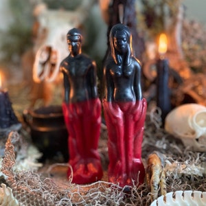 Könnte beinhalten: Zwei schwarze und rote Kerzenfiguren, eine männliche und eine weibliche, stehen auf einem Bett aus getrocknetem Moos und Knochen. Die Figuren sind in Rot gekleidet und haben einen schwarzen Oberkörper.