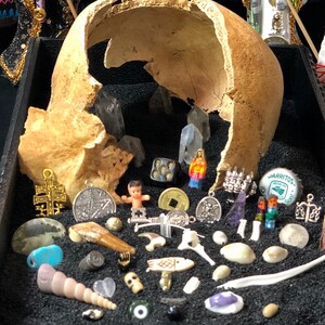 Può includere: Una collezione di piccoli oggetti decorativi disposti su una superficie nera all'interno di un cranio umano rotto. Gli oggetti includono una piccola bambola, un dado, una moneta, un cristallo, un tappo di bottiglia, una conchiglia, un piccolo teschio e una varietà di altri gingilli.