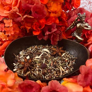May include: A dark, oval bowl filled with dried herbs and star anise, surrounded by vibrant red and orange flowers. A small bronze statue of a man is visible in the background, adding a touch of classical art to the composition.