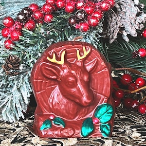 May include: A brown ceramic candle shaped like a deer head with gold antlers and green holly leaves. The candle is sitting on a gold and green patterned surface.