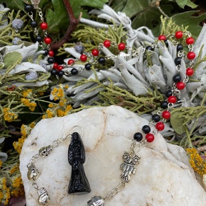 May include: A black and red beaded necklace with a silver charm of a woman in a black robe, a silver skull charm, and a silver owl charm.