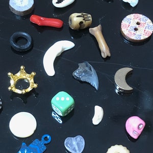 May include: A collection of small, colorful, and unique trinkets and charms, including a seashell, a bone, a skull, a crown, a dice, a hamsa hand, a heart, a moon, and a few other assorted items.