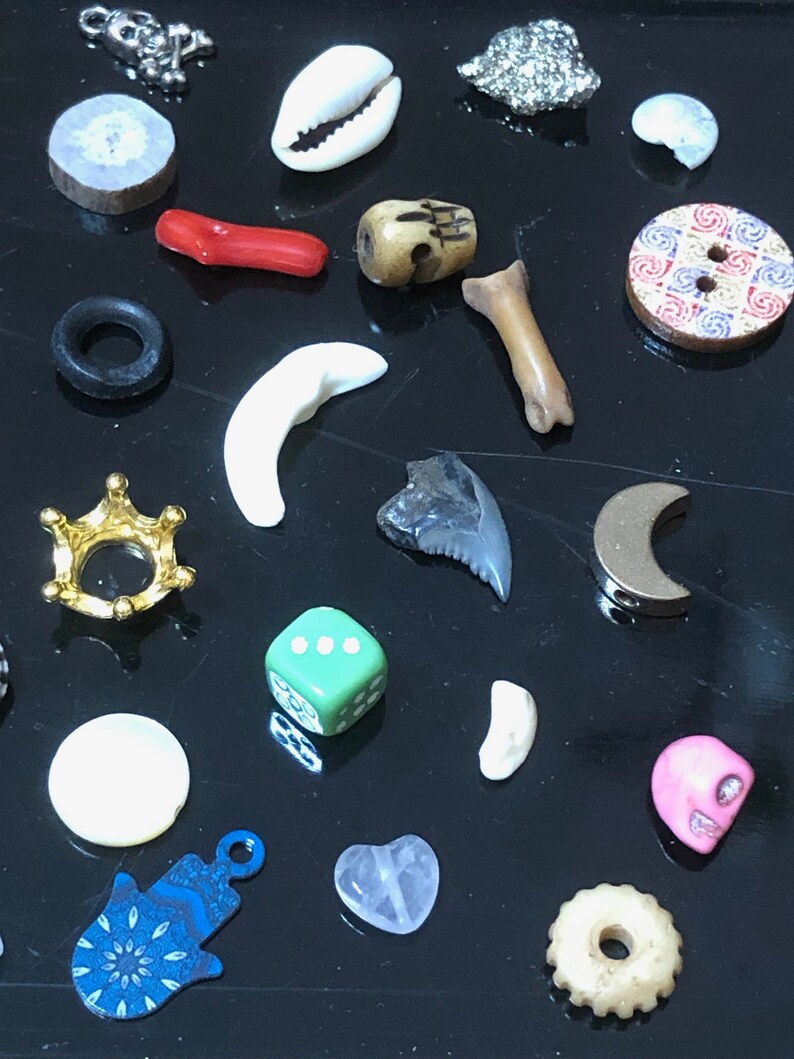 May include: A collection of small, colorful, and unique trinkets and charms, including a seashell, a bone, a skull, a crown, a dice, a hamsa hand, a heart, a moon, and a few other assorted items.