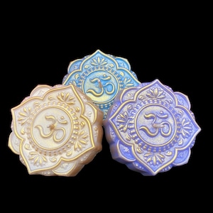 Puede incluir: Tres velas decorativas con forma de flor de loto con un símbolo Om en el centro. Las velas son de color azul, morado y blanco.