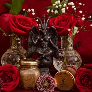 May include: A black Baphomet statue sits atop a globe, flanked by red roses in glass vases. A jar of brown powder, a purple flower, and a gold-lidded container of incense are in the foreground. The background is a deep red.