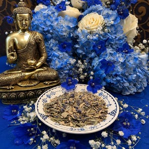 May include: A gold-toned Buddha statue in a seated position next to a white ceramic bowl filled with dried herbal tea leaves. The arrangement is surrounded by a vibrant bouquet of blue delphiniums, white roses, and small white baby's breath flowers.