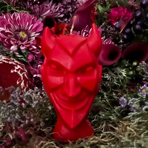 Puede incluir: Una vela roja con forma de cabeza de diablo con cuernos, sobre un fondo de flores moradas y rojas.