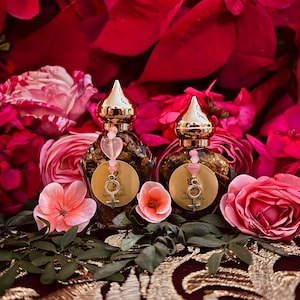 May include: Two small glass bottles filled with herbs and crystals. The bottles have gold tops and are labelled "Divine Feminine Spiritual Arts".