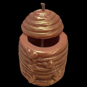 May include: A pink and gold honeycomb-shaped candle with a lid. The candle is designed to resemble a beehive.