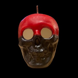 May include: A red and black skull-shaped candle with a wick. The top half of the skull is red and the bottom half is black.