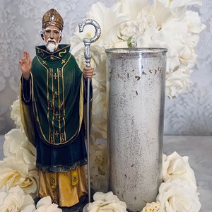 以下が含まれることがあります： 金色のグリッターデザインが施されたガラスの瓶に入った白いキャンドル。キャンドルは白い花と、緑色のローブと金色のミトラを身に着けた聖人の像に囲まれています。