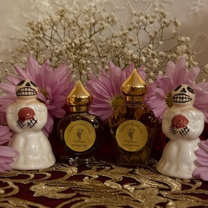 Könnte beinhalten: Vier Parfümflaschen mit goldenen Verschlüssen und Etiketten mit der Aufschrift "Lesbian Marriage Serpentine Spiritual Arts" werden zusammen mit zwei weißen Keramikfiguren mit Totenkopfgesichtern und roten Akzenten präsentiert. Rosa und weiße Blumen sind im Hintergrund.