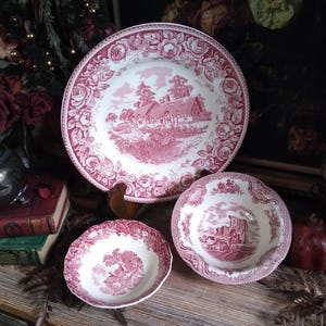 May include: A set of antique red and white ceramic dishes. The set includes a large plate and two smaller bowls, all with a detailed landscape scene and floral border. The dishes are arranged on a wooden surface.