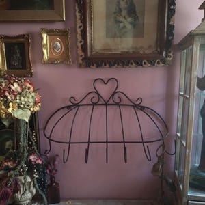 May include: Black metal wall-mounted rack with a heart-shaped design at the top. The rack has a curved, cage-like structure with several hooks below. The background features framed artwork and a pink wall.