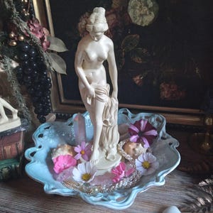 May include: A cream-colored statue of a woman holding a draped cloth, standing in a light blue, scalloped-edge dish. The dish contains pink and white flowers, crystals, and jewelry. A dark floral painting is in the background.