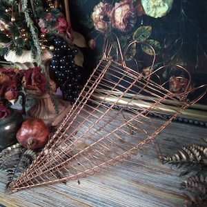 May include: A copper-colored, cone-shaped wire basket with a heart-shaped handle. The basket is made of thin, interwoven wires, creating a lattice pattern. The background includes a still-life painting of flowers and fruit.