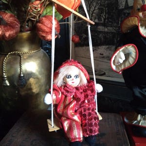 May include: A vintage doll in a red and gold costume with a hood, seated on a swing made of white rope and a wooden seat. The doll has a painted face and is part of a decorative display. A clown doll is in the background.