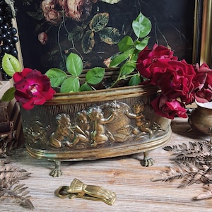May include: An oval brass planter with four claw feet, featuring a relief design of cherubs playing with a globe. Deep red roses with green leaves are placed inside the planter. A brass clip shaped like a hand rests in front of the planter.