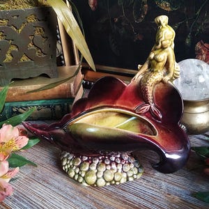 以下が含まれることがあります： 貝殻型の皿に乗った、ヴィンテージのセラミック人魚の置物。皿は、ブルゴーニュ、黄色、緑色のグラデーションで、テクスチャード加工されたベースが付いています。金色の人魚が、この装飾品に気まぐれなタッチを加えています。