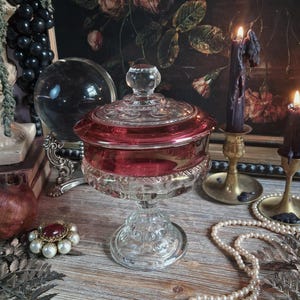 May include: A clear and cranberry red glass candy dish with a lid, featuring a pedestal base and intricate pressed glass patterns. The dish has a wide bowl with a deep red band and a matching lid with a small knob handle, standing on a textured stem.